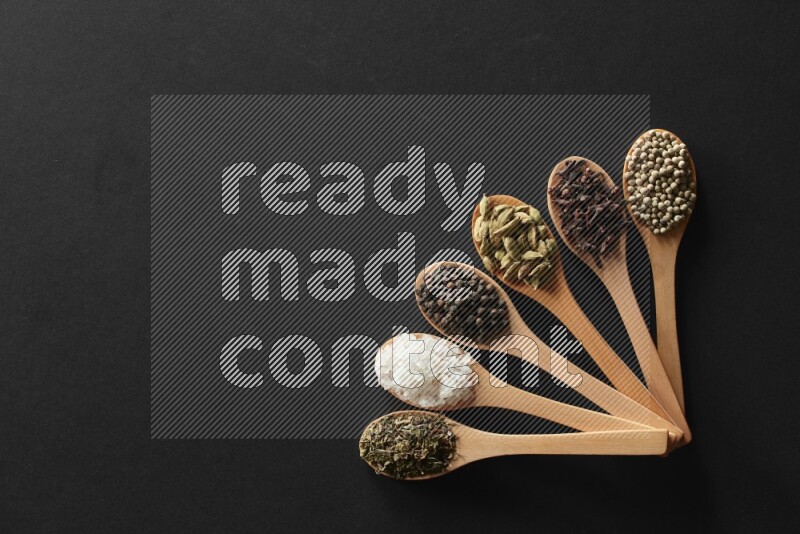 wooden spoons filled with white peppers, cloves, cardamom, salt, black peppers and basil on black flooring