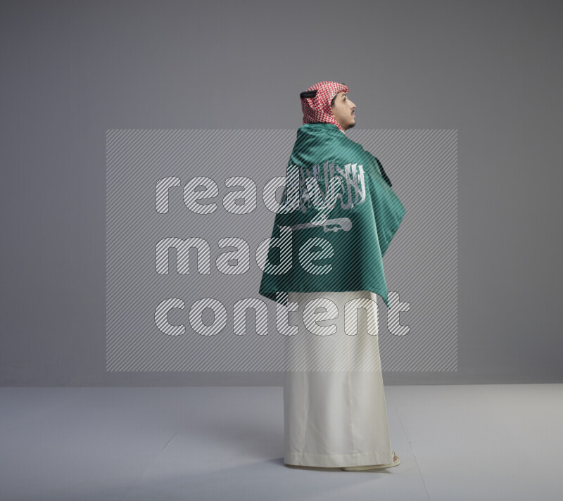 A Saudi man standing wearing thob and red shomag wrapping big saudi flag on gray background