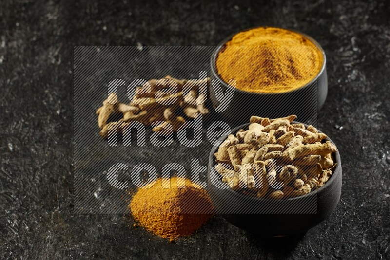2 pottery black bowls, one full of turmeric powder and the other full of dried turmeric whole fingers on a textured black flooring