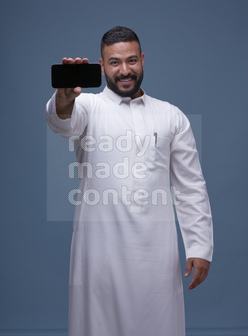 A man Showing his phone in Blue Background wearing Saudi Thob
