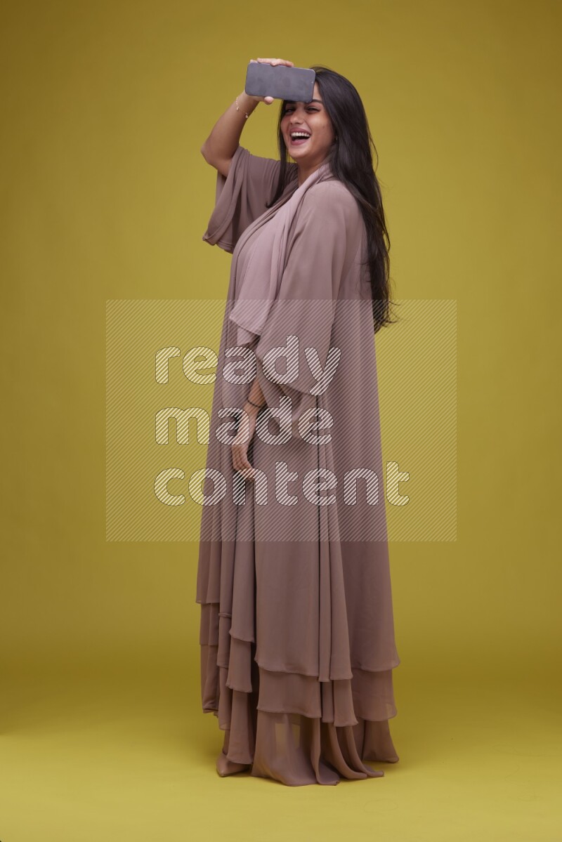 A woman Showing SmartPhone Screen on a Yellow Background wearing Brown Abaya