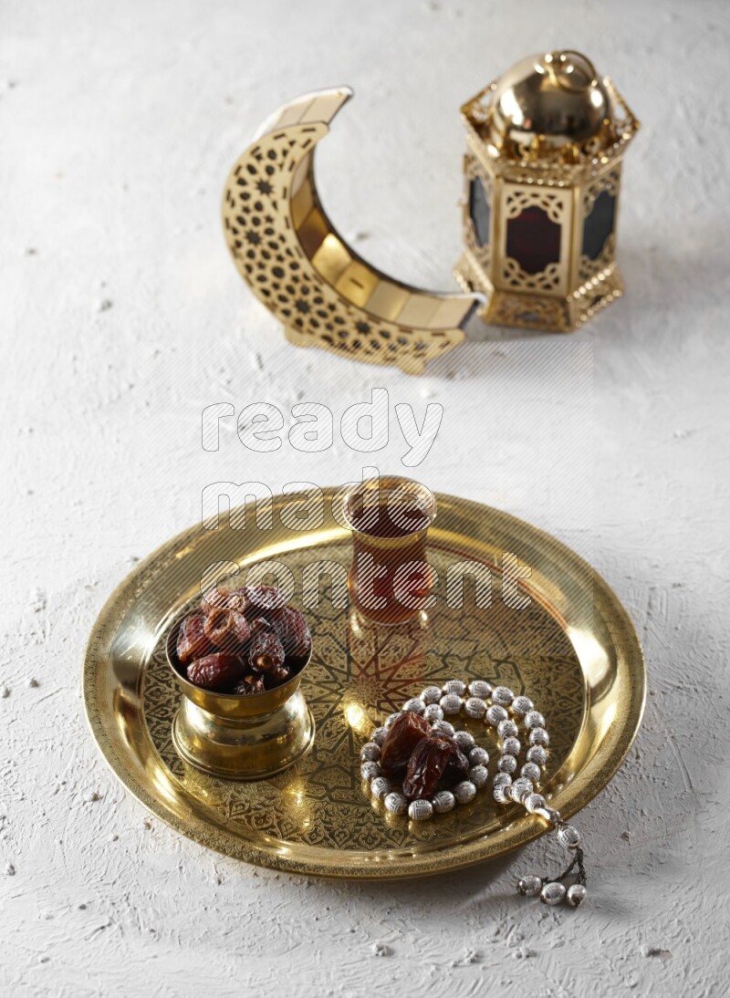 Dates in a metal bowl with tea and prayer beads on a tray beside lanterns in a light setup