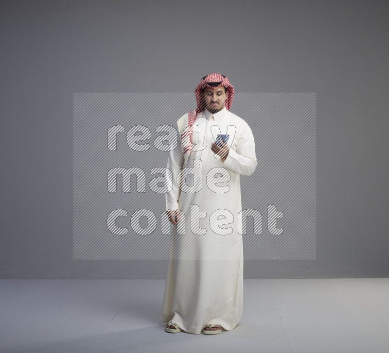 A  Saudi man standing wearing thob and red shomag texting on phone on gray background