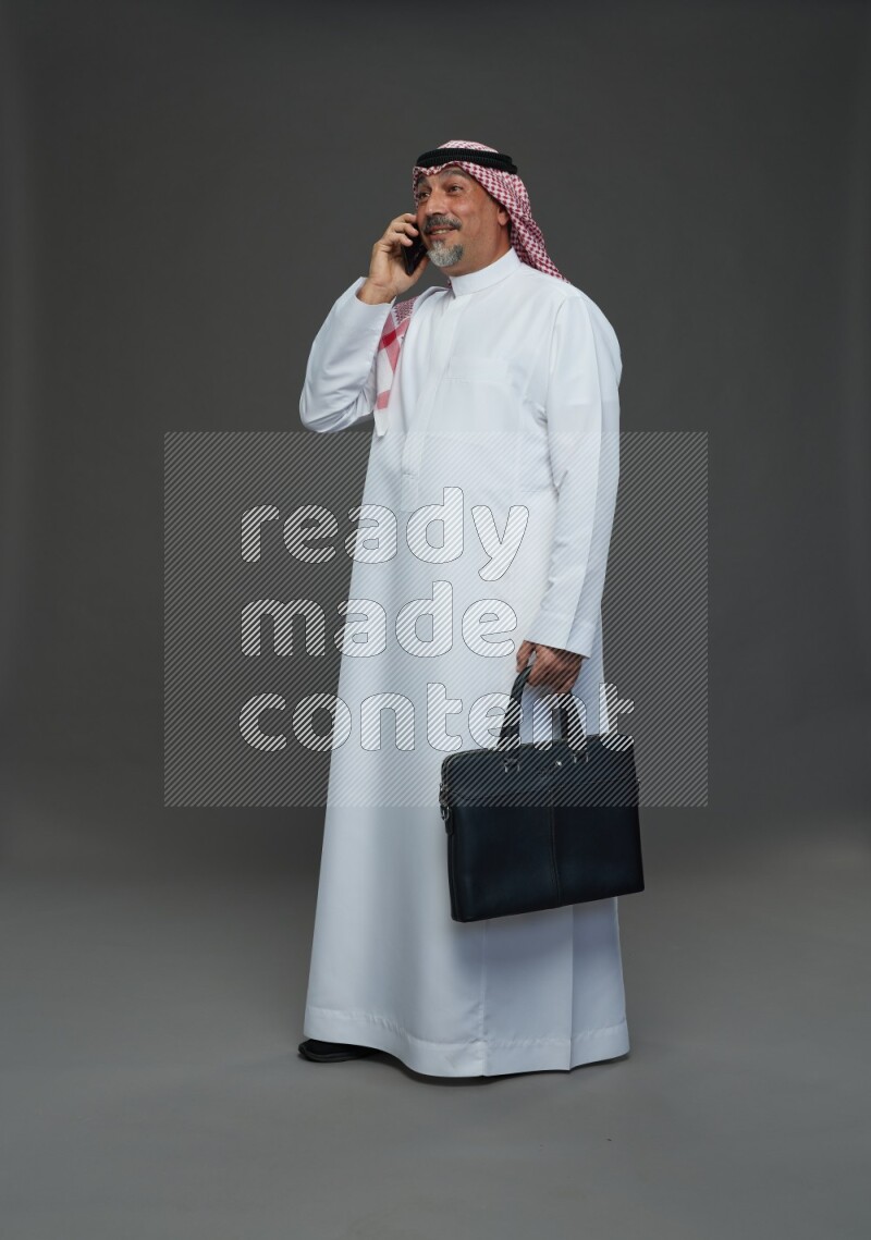 Saudi man with shomag Standing holding bag talking on phone on gray background