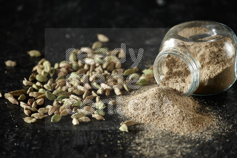 A flipped glass spice jar full of cardamom powder and powder spilled out of it with cardamom seeds spreaded on textured black flooring