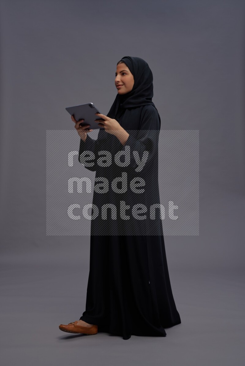 Saudi woman wearing Abaya standing working on tablet on gray background