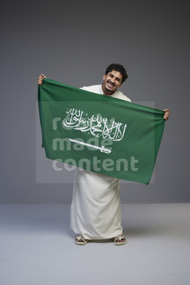 A Saudi man standing wearing thob holding big Saudi flag on gray background