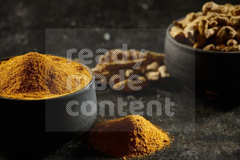 2 pottery black bowls, one full of turmeric powder and the other full of dried turmeric whole fingers on a textured black flooring