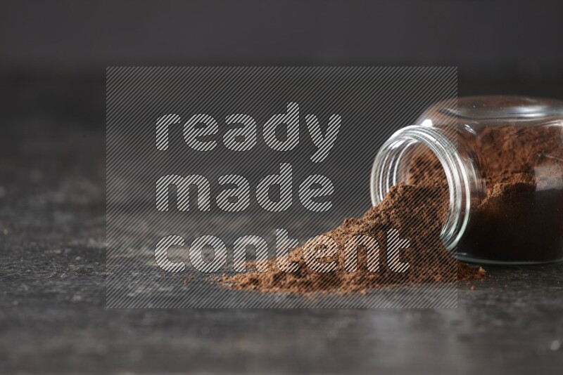 A flipped glass spice jar full of cloves powder and powder came out of it on textured black flooring