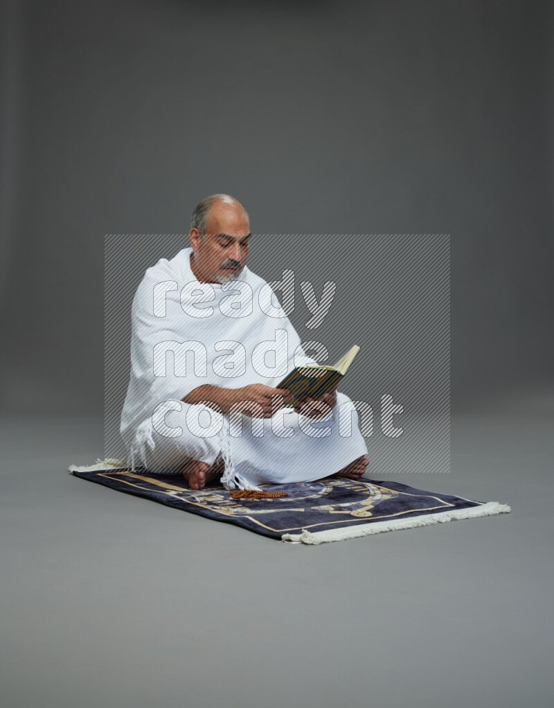 A man wearing Ehram sitting on mate prayer reading quran on gray background