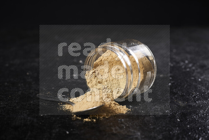 A glass jar full of ground ginger powder flipped with some spilling powder on black background