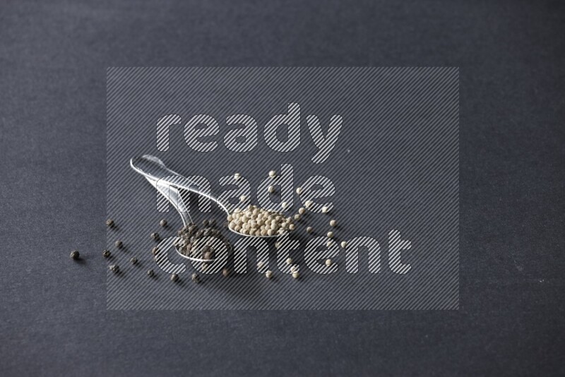 2 metal spoons full of black and white pepper beads with spreaded beads on black flooring