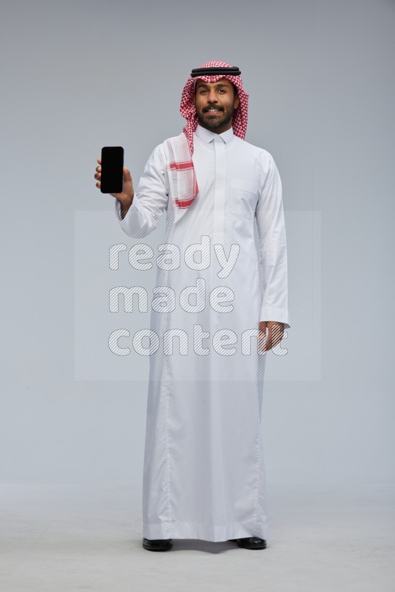 Saudi man Wearing Thob and shomag standing showing phone to camera on Gray background