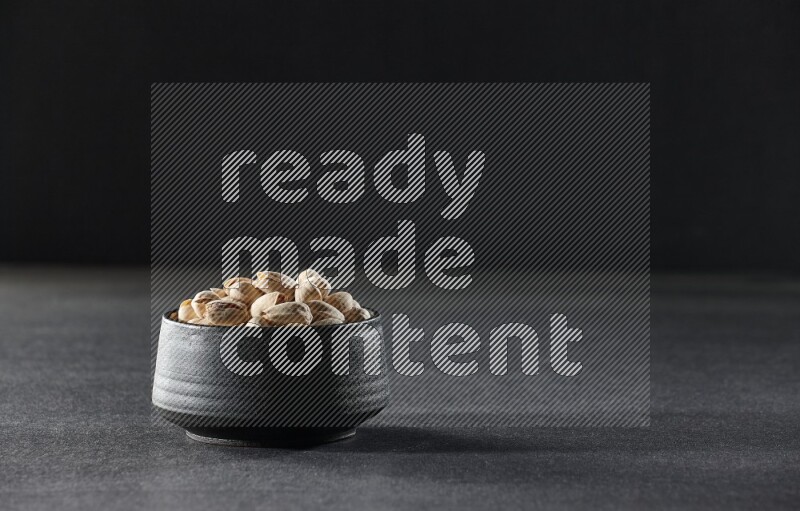A black pottery bowl full of pistachios on a black background in different angles