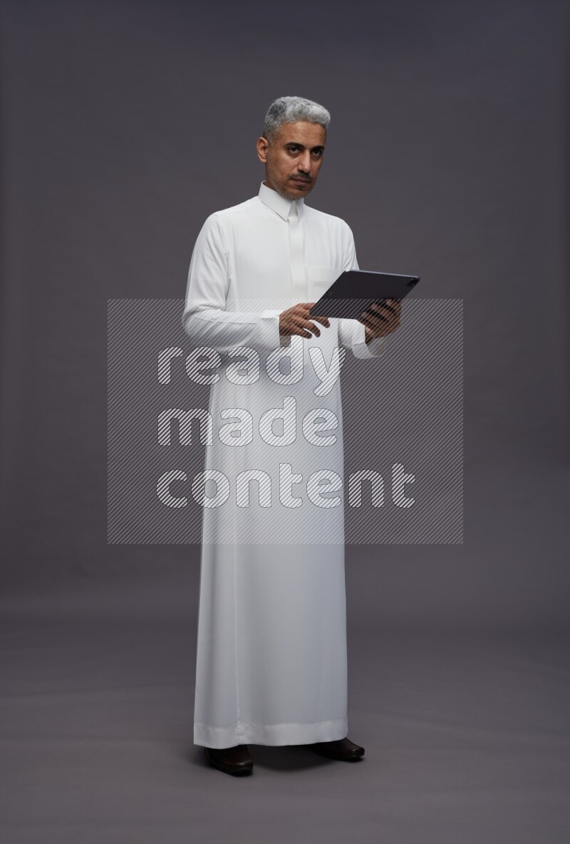 Saudi man wearing thob standing working on tablet on gray background