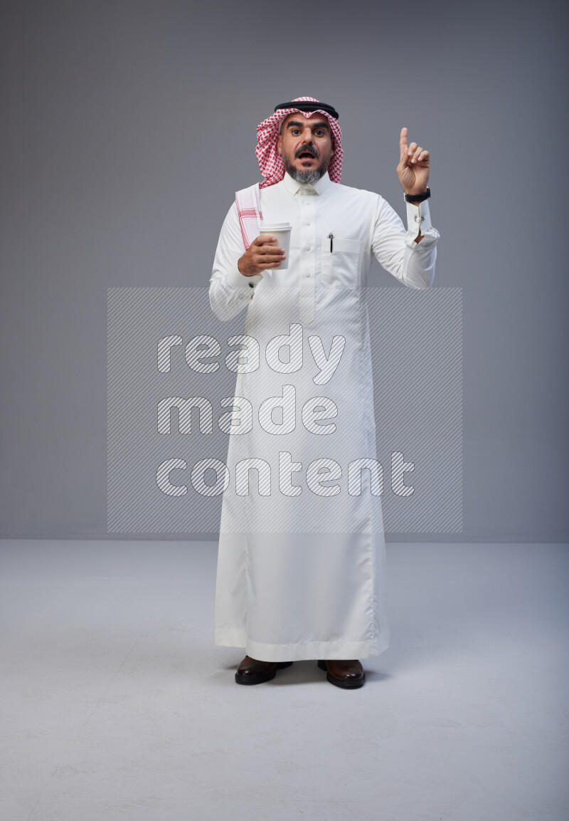 Saudi man Wearing Thob and red Shomag standing holding paper cup on Gray background