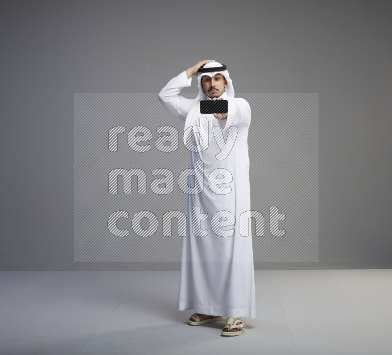 A Saudi man standing wearing thob and white shomag showing phone to camera on gray background