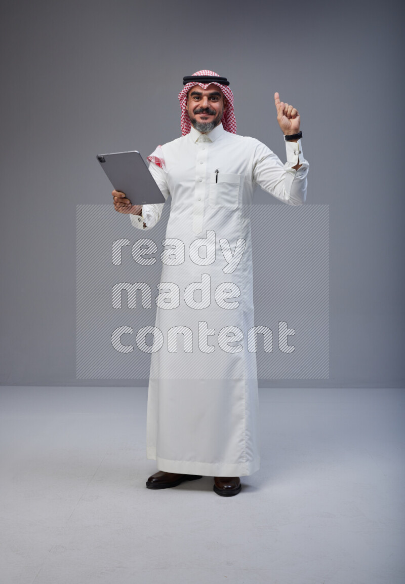 Saudi man Wearing Thob and red Shomag standing working on tablet on Gray background