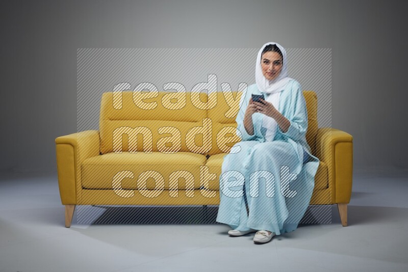 A Saudi woman wearing a light blue Abaya and white head scarf sitting on a yellow sofa and texting on her phone on a grey background