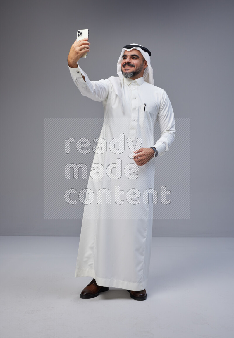 Saudi man Wearing Thob and white Shomag standing taking selfie on Gray background