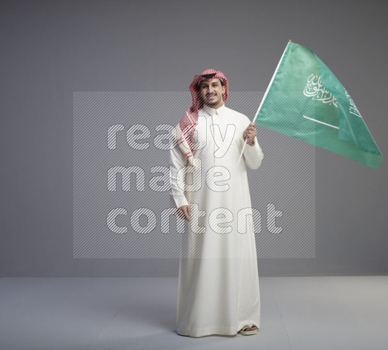 A Saudi man standing wearing thob and red shomag raising big Saudi flag on gray background