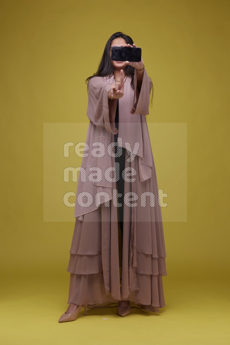 A woman Showing SmartPhone Screen on a Yellow Background wearing Brown Abaya