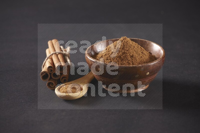Cinnamon sticks stacked and bounded beside a wooden bowl full of cinnamon powder and a wooden spoon full of powder on black background