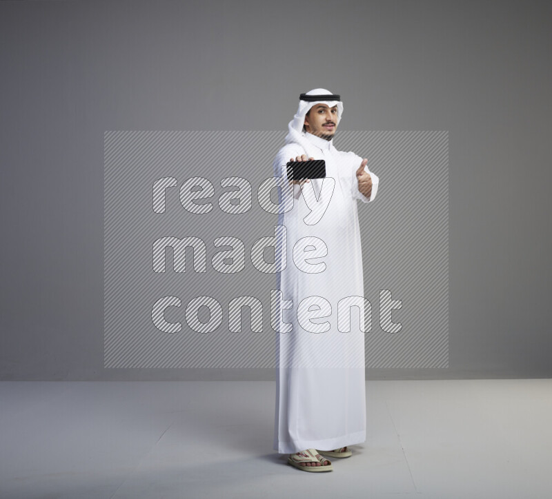 A Saudi man standing wearing thob and white shomag showing phone to camera on gray background