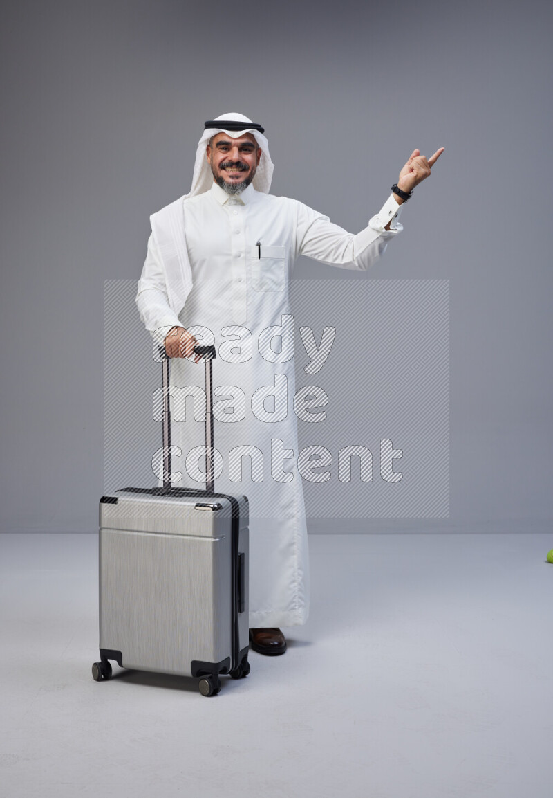 Saudi man wearing Thob and white Shomag standing holding Travel bag on Gray background
