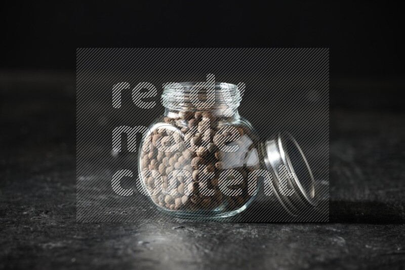 A glass spice jar full of allspice whole balls on a textured black flooring