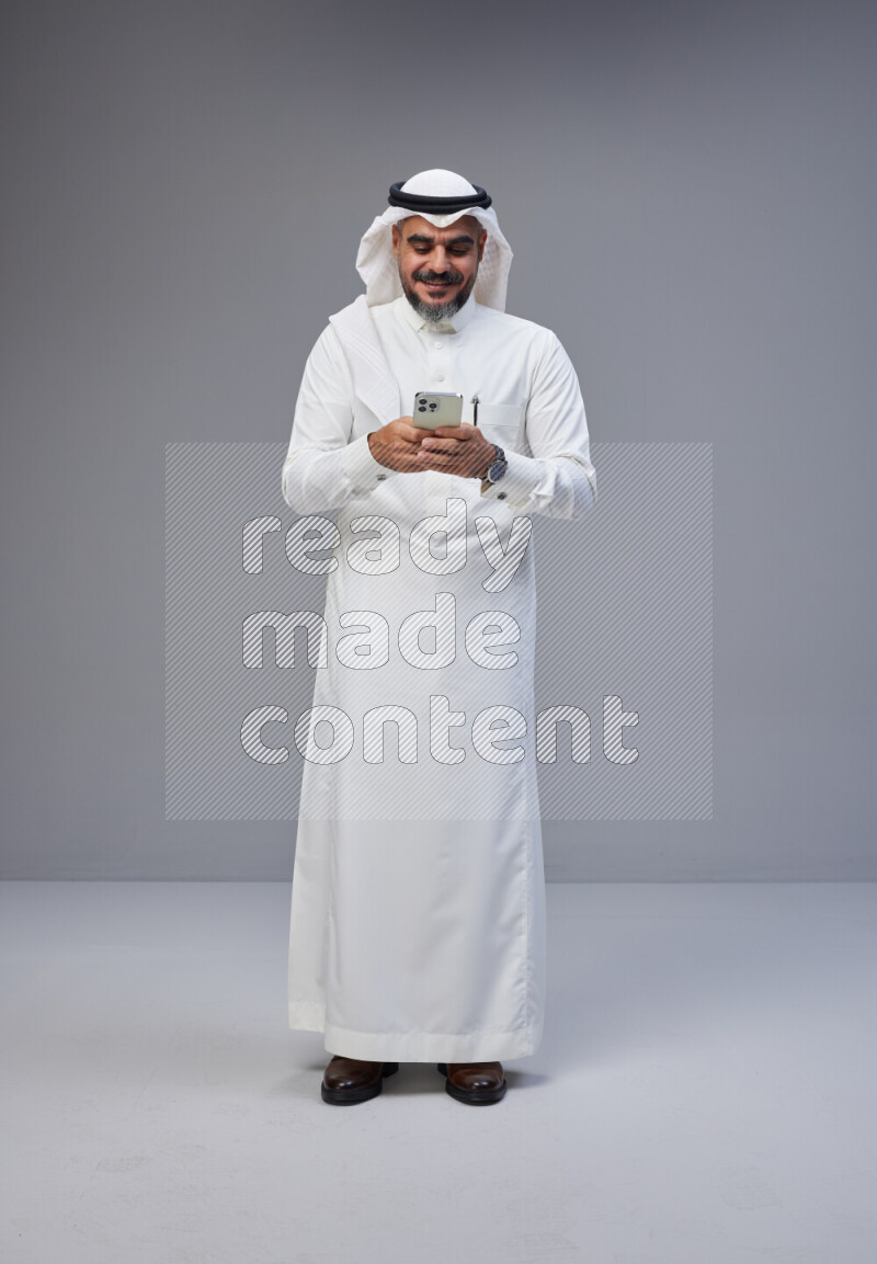 Saudi man Wearing Thob and white Shomag standing texting on phone on Gray background