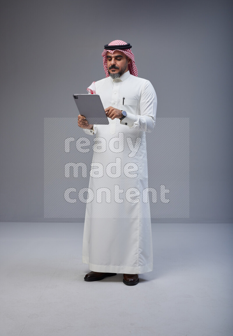 Saudi man Wearing Thob and red Shomag standing working on tablet on Gray background