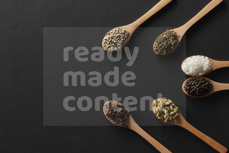 wooden spoons filled with white peppers, cloves, cardamom, salt, black peppers and basil on black flooring