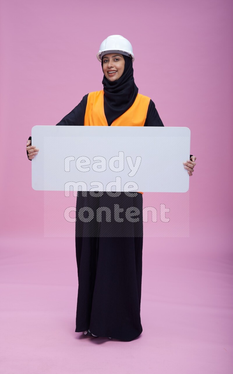 Saudi woman wearing Abaya with engineer vest and helmet standing holding board on pink background