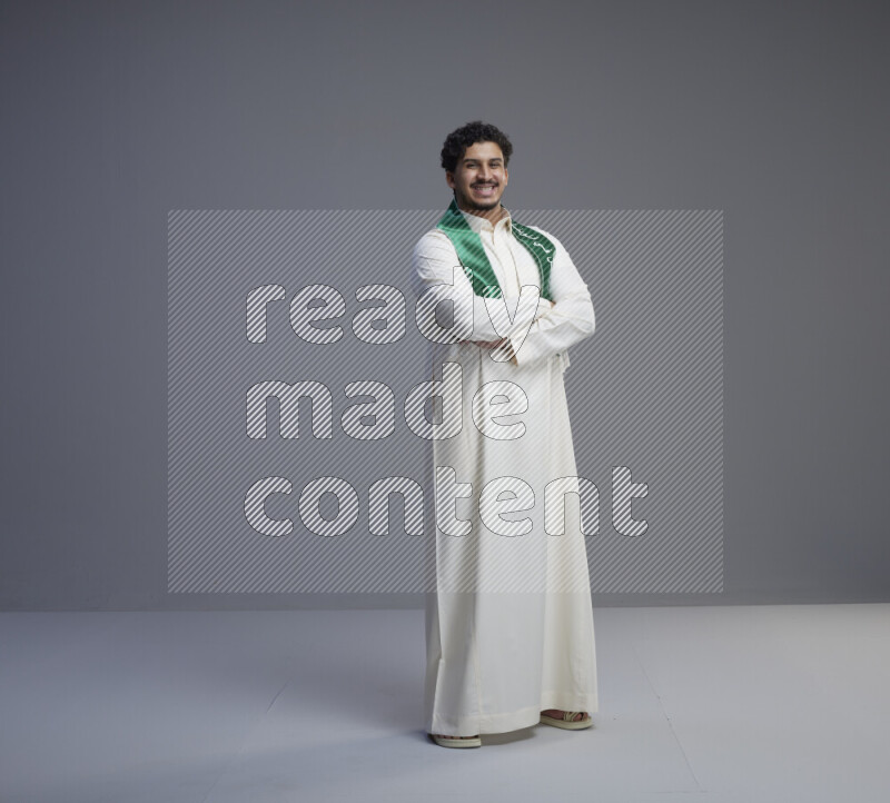A Saudi man standing wearing thob and Saudi flag scarf on gray background