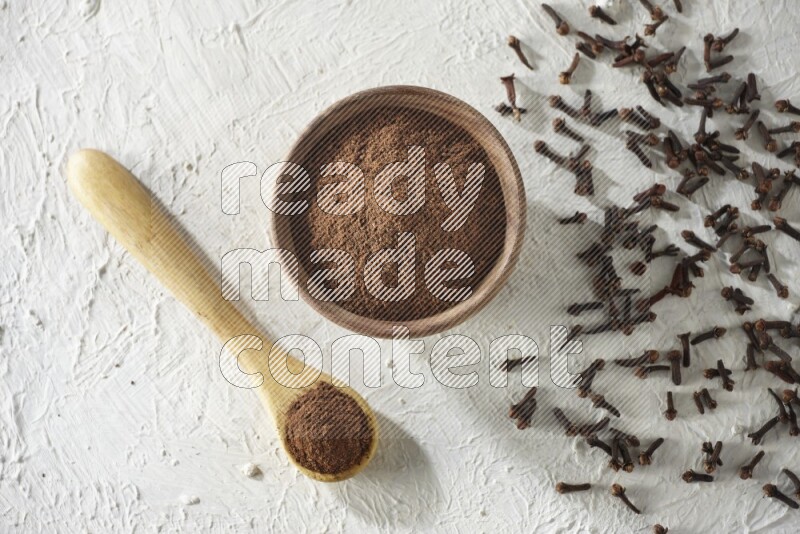 A wooden bowl and wooden spoon full of cloves powder with cloves spread on textured white flooring