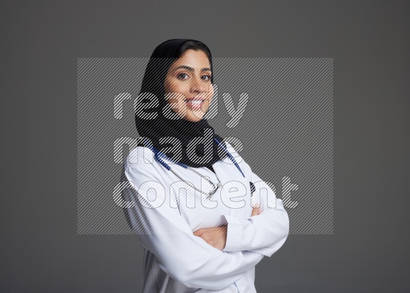Saudi woman wearing lab coat with stethoscope standing with crossed arms on Gray background