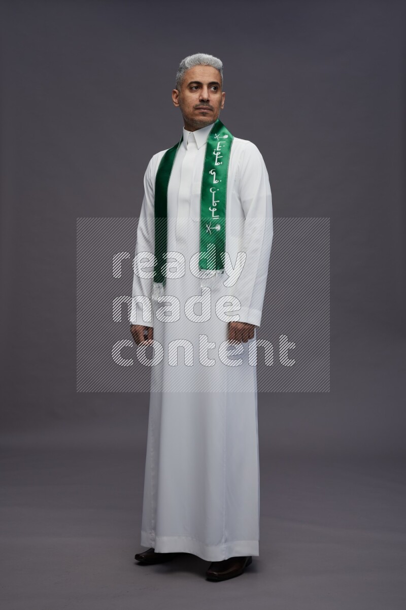 Saudi man wearing thob standing holding Saudi flag on gray background