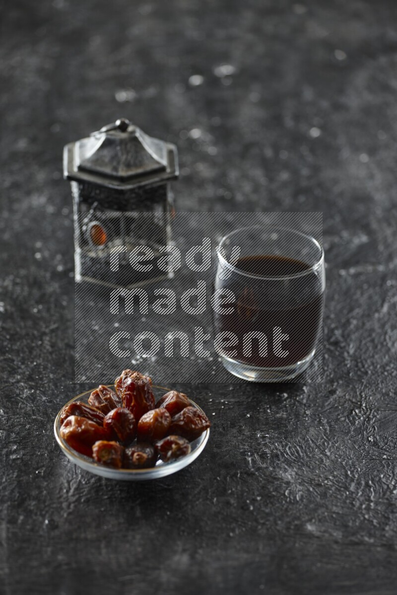 A silver lantern with different drinks, dates, nuts, prayer beads and quran on textured black background
