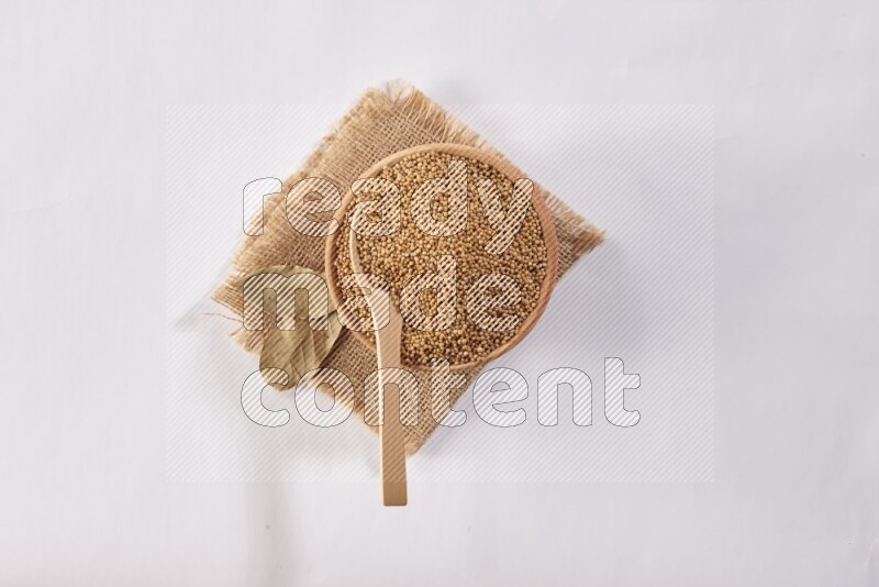 A wooden bowl full of mustard seeds with a wooden spoon on the seed set on burlap fabric on a white flooring