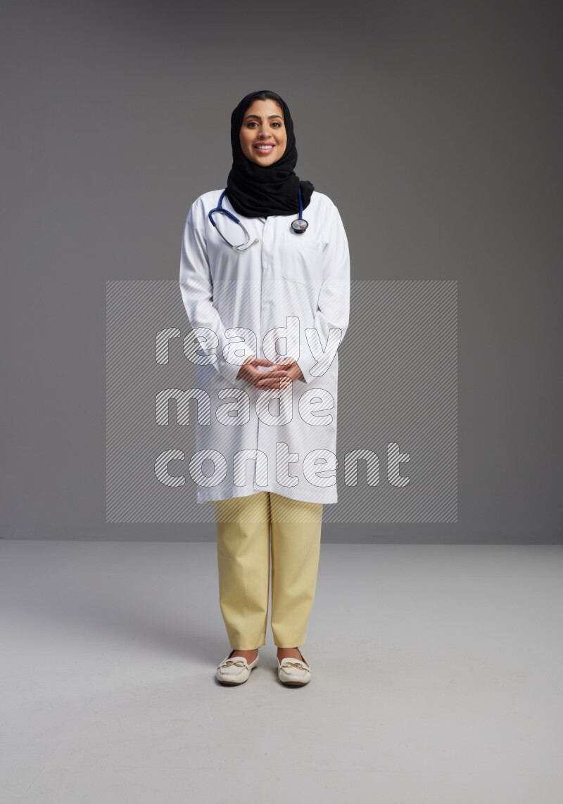 Saudi woman wearing lab coat with stethoscope standing interacting with the camera on Gray background
