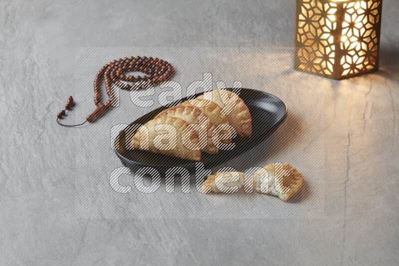 Four fried sambosas in an oval shaped black plate, beside a cut cheese sambosa, a brown misbaha and a golden lantern on a gray background