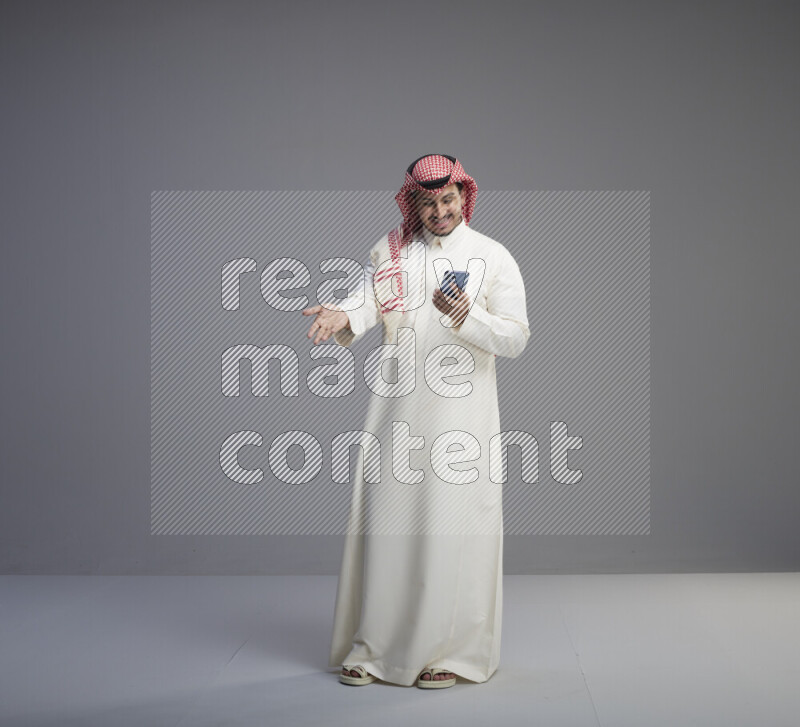 A  Saudi man standing wearing thob and red shomag texting on phone on gray background