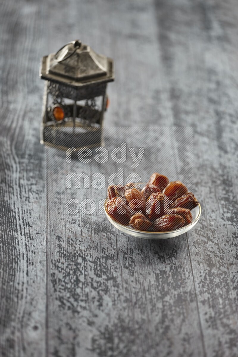 A silver lantern with different drinks, dates, nuts, prayer beads and quran on grey wooden background