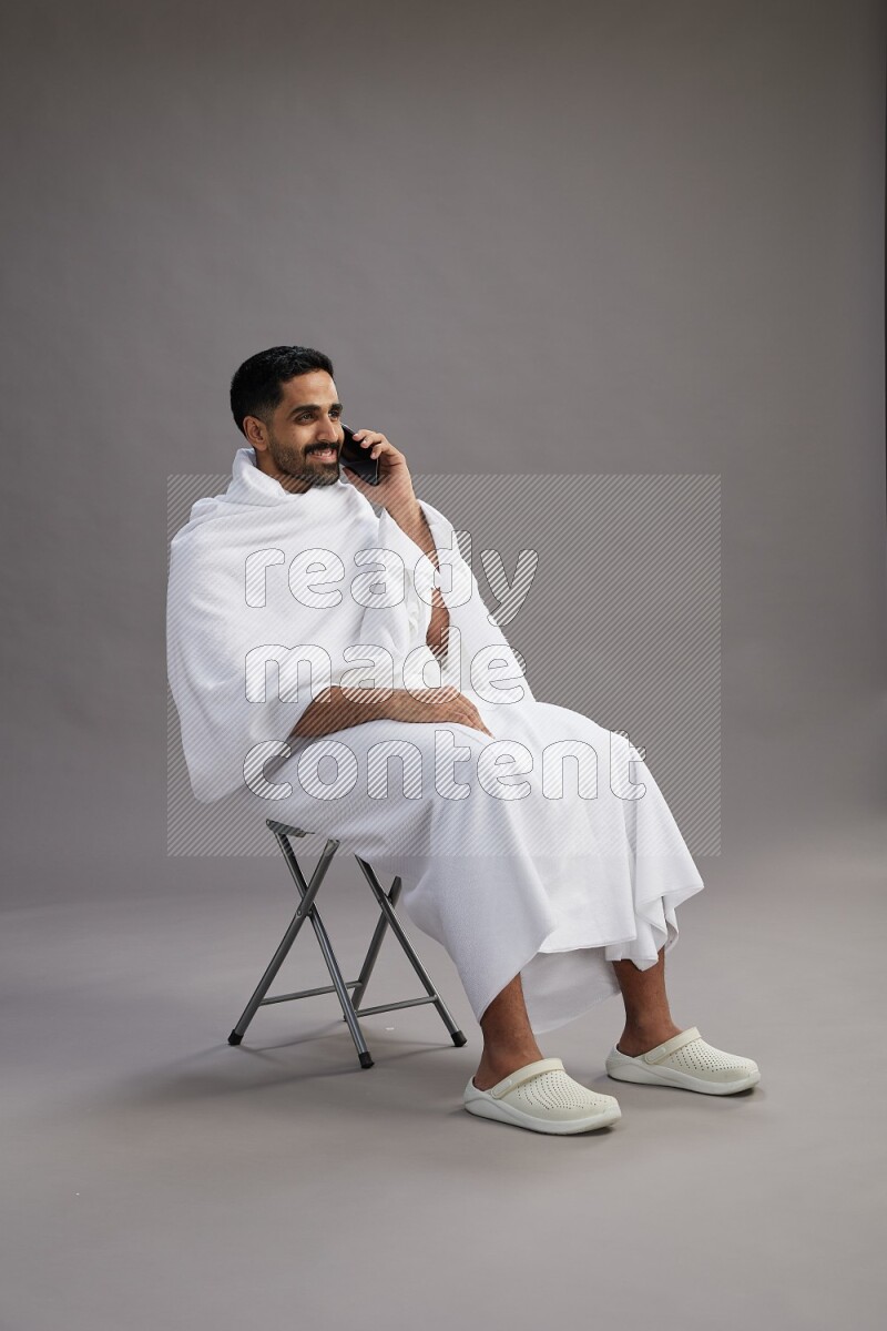 A man wearing Ehram sitting on chair talking on the phone on gray background