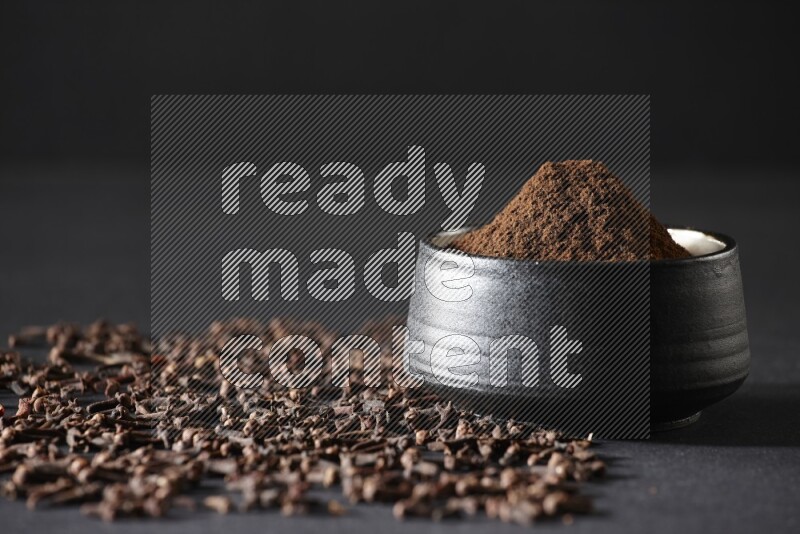 وعاء فخاري أسود ممتلئ ببودرة القرنفل وحبوب القرنفل على خلفية سوداء