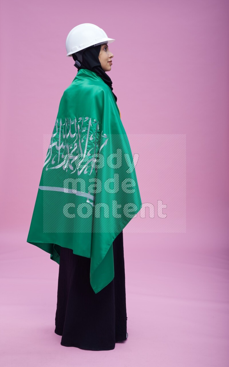 Saudi woman wearing Abaya with engineer vest and helmet standing holding Saudi flag on pink background