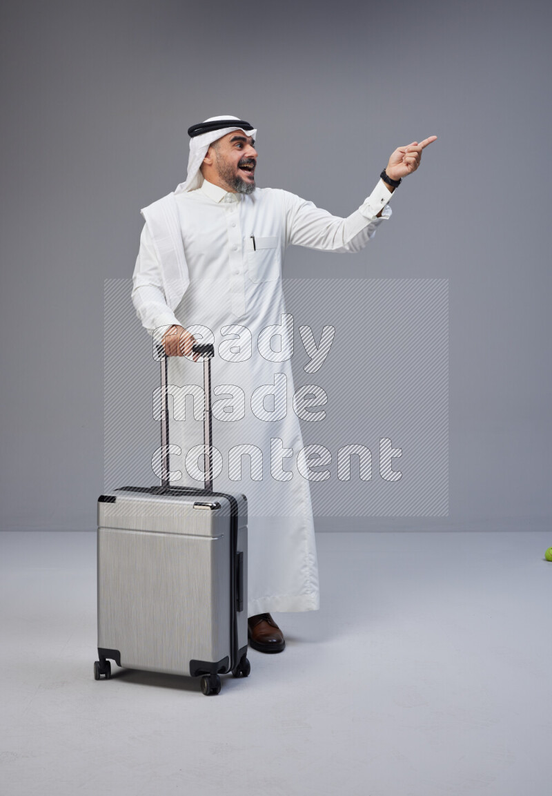 Saudi man wearing Thob and white Shomag standing holding Travel bag on Gray background