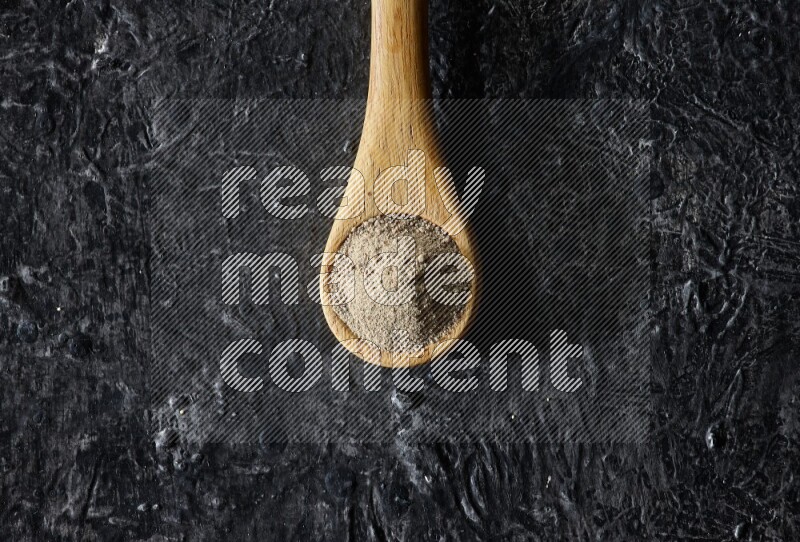 A wooden spoon full of white pepper powder on textured black flooring