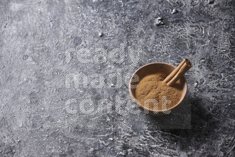 Wooden bowl full of cinnamon powder and a cinnamon stick on a textured black background